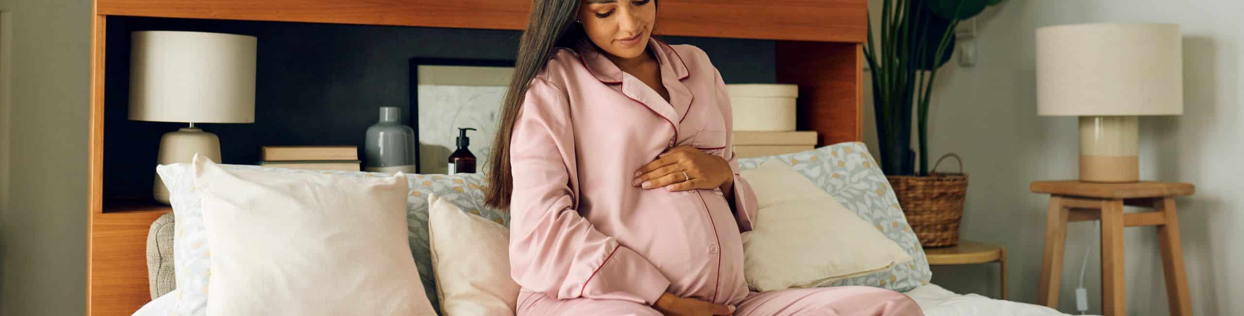 Trudnica sjedi u krevetu i dodiruje svoj trbuh - važnost vitamina u trudnoći